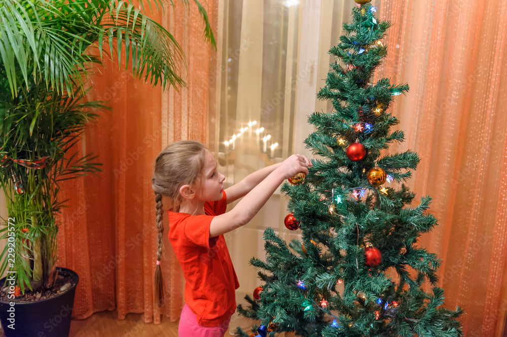 Adorable little girl decorating a Christmas tree with colorful glass baubles at home