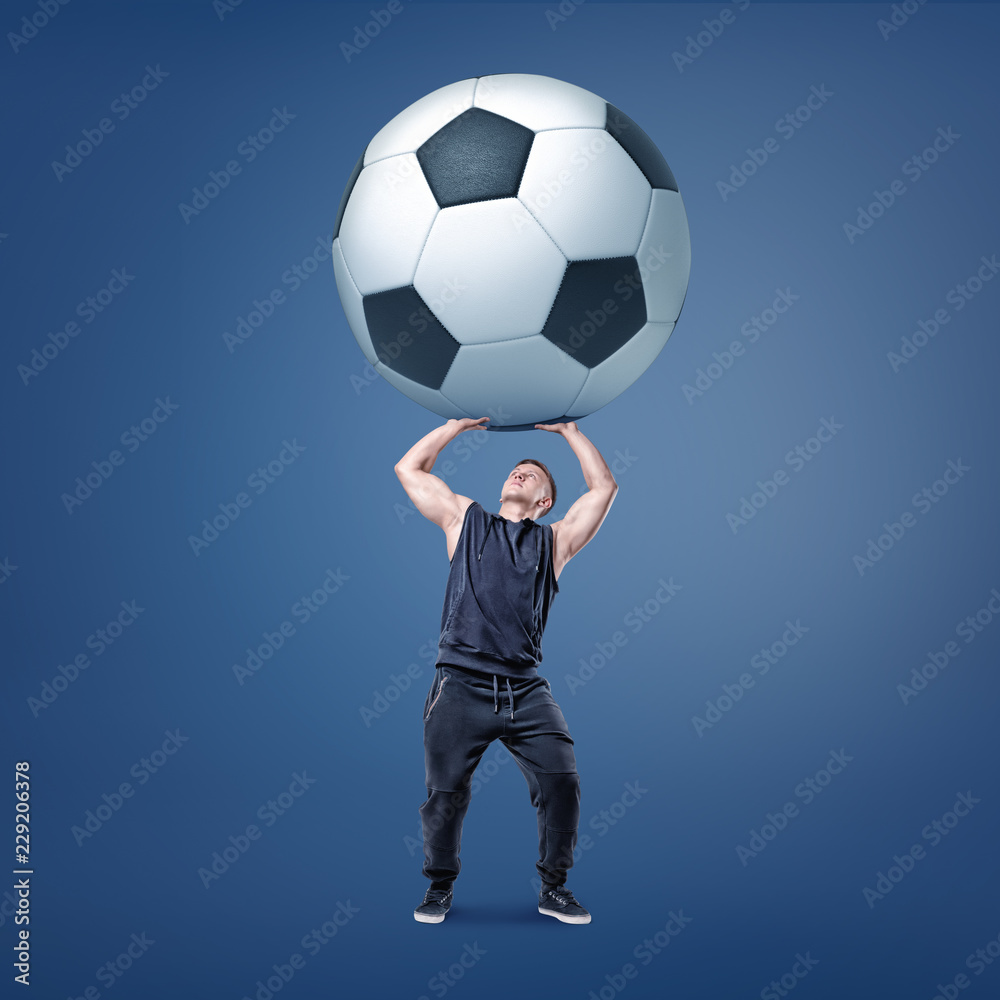A muscular man tries to hold a giant and heavy black and white football ball over himself.