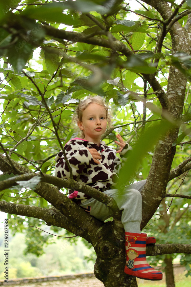 Little curly girl in a spotted fleece and rubber boots is sitting on a tree. Vacation in the village, happy childhood Little curly girl in a spotted fleece and rubber boots is sitting on a tree. Vacation in the village, happy childhood