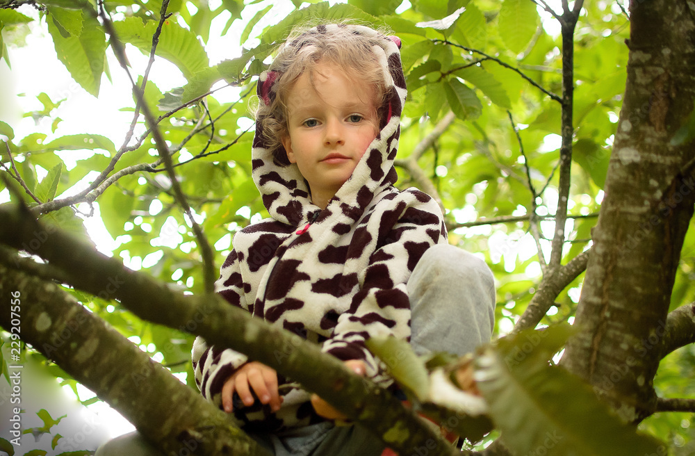 Little curly girl in a spotted fleece and rubber boots is sitting on a tree. Vacation in the village, physical activity Little curly girl in a spotted fleece and rubber boots is sitting on a tree. Vacation in the village, physical activity