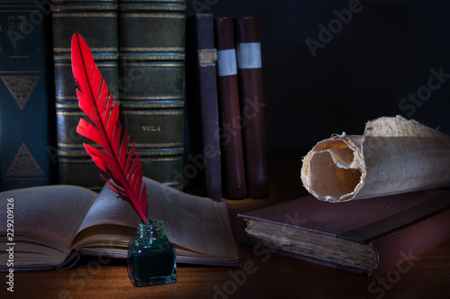 Red quill pen and a rolled papyrus sheet on a wooden table with old books