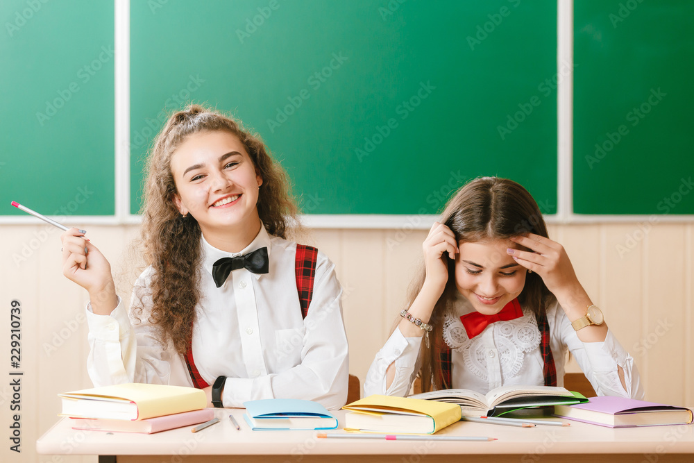 2 schoolgirls in school uniform sit on the background of a green board at the desk with books 2 schoolgirls in school uniform sit on the background of a green board at the desk with books