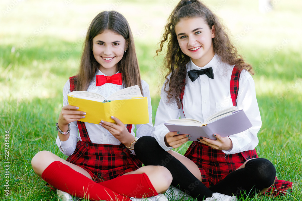 Fototapeta premium two funny schoolgirls sit on the grass and read books. Girls, girlfriends, sisters are taught lessons in nature