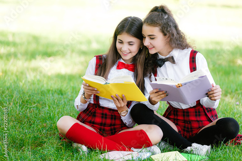two funny schoolgirls sit on the grass and read books. Girls, girlfriends, sisters are taught lessons in nature