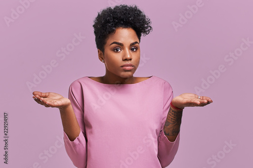 Fotografie Uncertain African American female with dark skin, shruggs shoulders and asks something, has clueless expression, isolated on lavender background
