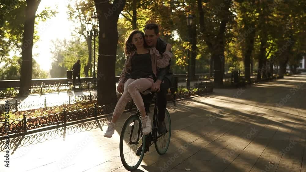 Smiling young couple riding on the same black bike with sun backlight on the background. Girl is sitting on the rudder while her boyfriend cycling. Front view Smiling young couple riding on the same black bike with sun backlight on the background. Girl is sitting on the rudder while her boyfriend cycling. Front view
