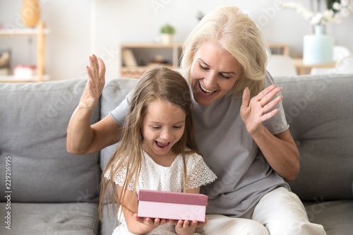 Smiling grandmother making surprise to cute little granddaughter giving gift box, excited small girl get birthday present from granny, happy older and younger generation celebrate at home together