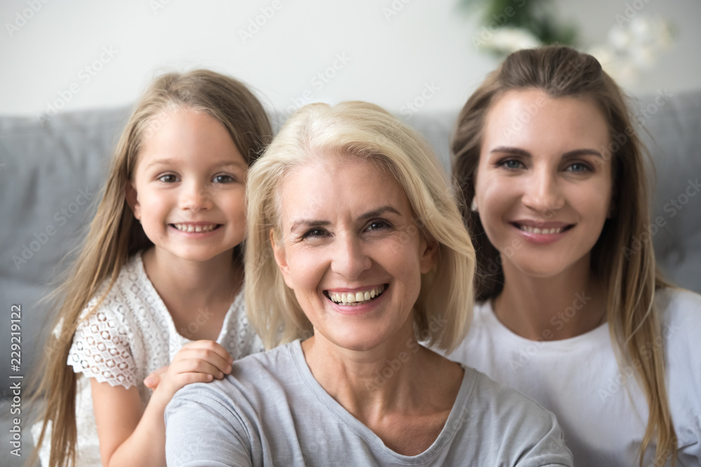 Portrait of smiling three generations of women look at camera posing for family picture at home, happy mother, daughter and grandmother in the middle embrace together showing love and support Portrait of smiling three generations of women look at camera posing for family picture at home, happy mother, daughter and grandmother in the middle embrace together showing love and support