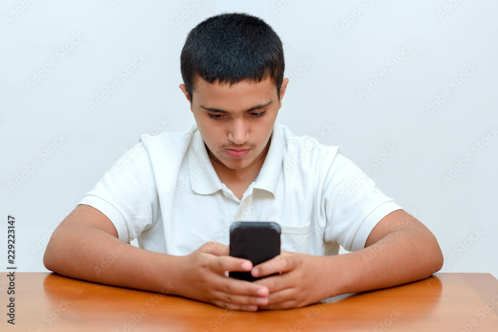Sitting young adult brunette boy writes a message on smartphone. Teenager with phone in hand sitting at table and studying at home. Sitting young adult brunette boy writes a message on smartphone. Teenager with phone in hand sitting at table and studying at home.