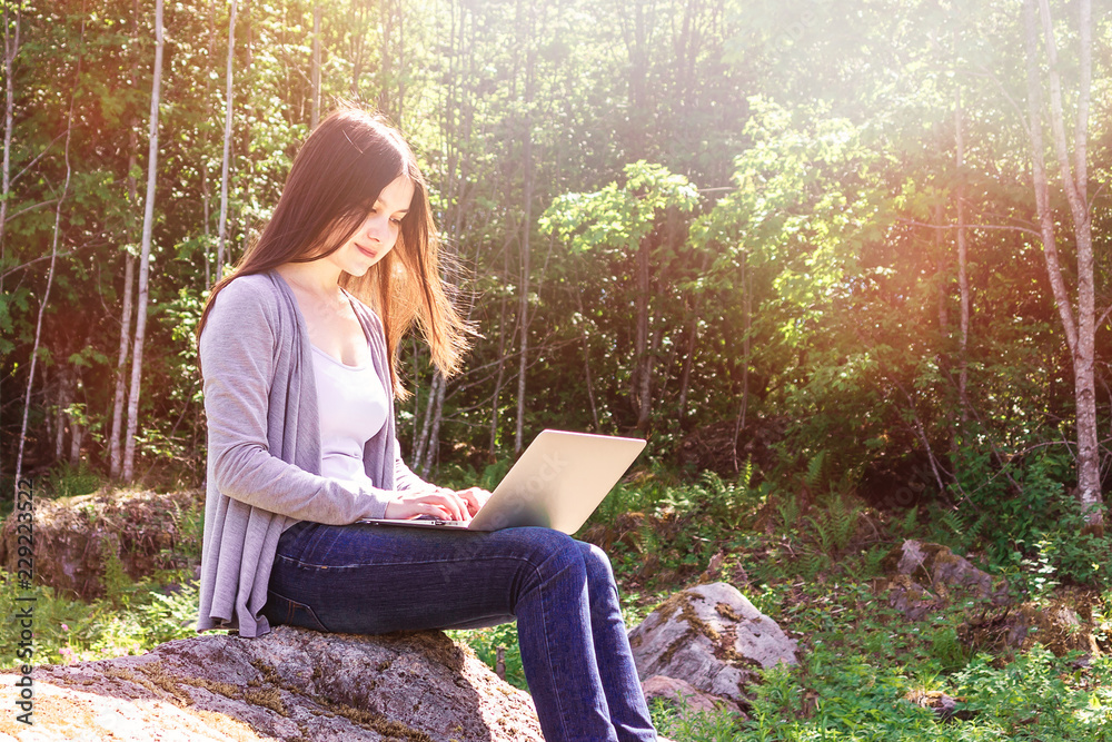 Young beautiful girl is working on a laptop while sitting on a big rock in the forest. Freelance concept Young beautiful girl is working on a laptop while sitting on a big rock in the forest. Freelance concept