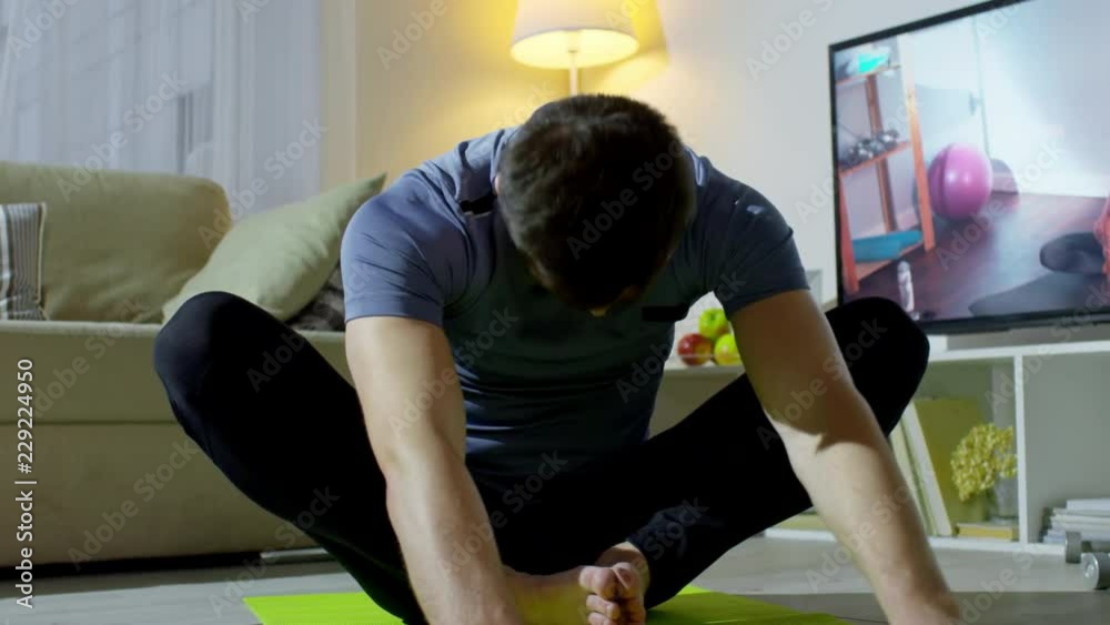 Medium shot of bearded man in sportswear sitting in lotus pose and bending forward to stretch, then resting and breathing hard while talking to female online personal trainer on TV screen Medium shot of bearded man in sportswear sitting in lotus pose and bending forward to stretch, then resting and breathing hard while talking to female online personal trainer on TV screen