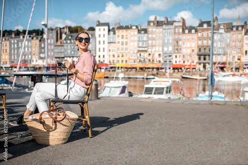 Young woman enjoying coffee sitting at the cafe outdoors near the harbour with beautiful buildings on the background in Honfleur old town, France