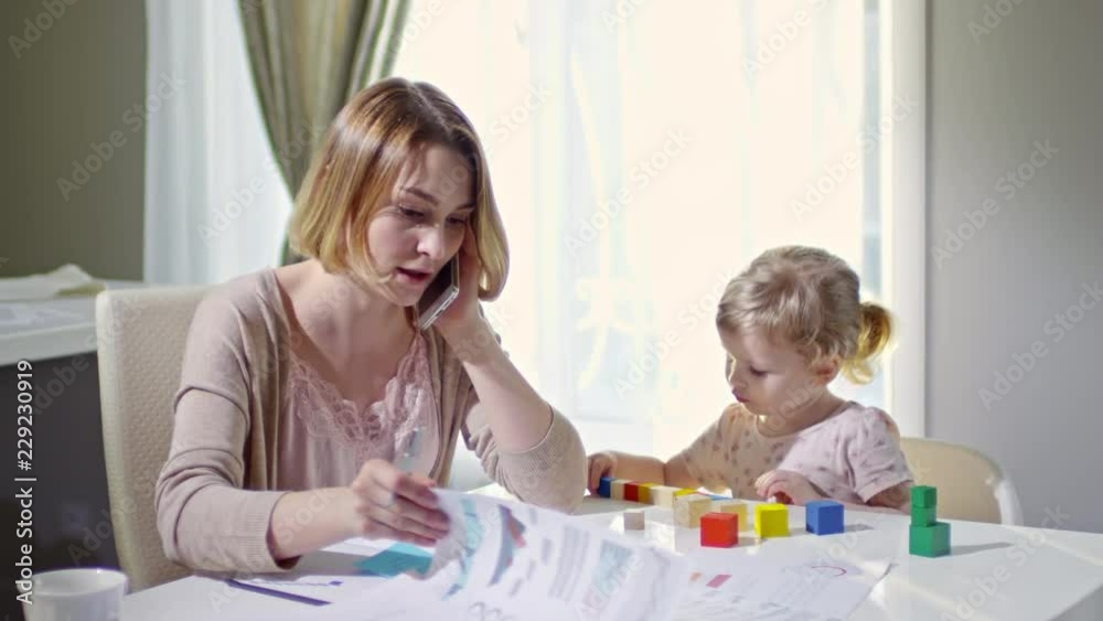 PAN of busy woman looking at document and talking on phone, then writing in her planner while working from home; cute toddler girl sitting beside her and playing with building blocks PAN of busy woman looking at document and talking on phone, then writing in her planner while working from home; cute toddler girl sitting beside her and playing with building blocks
