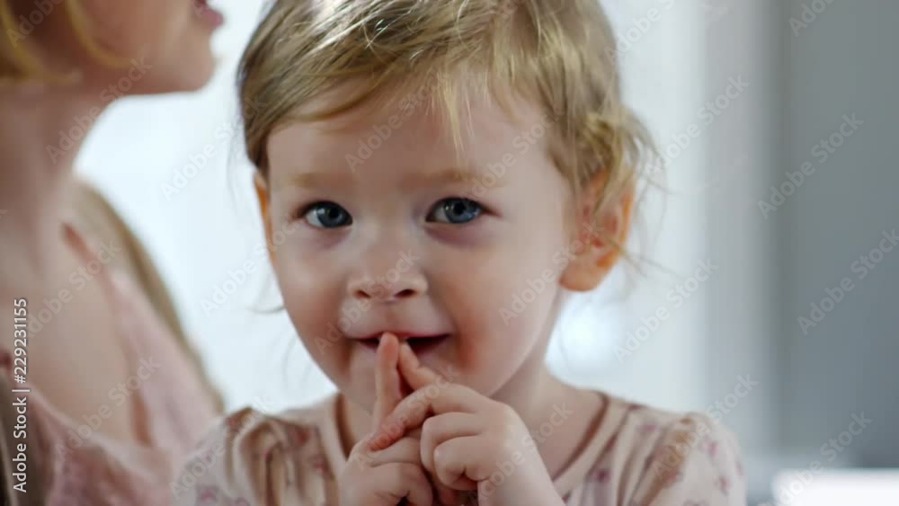 Close up of cute toddler girl smiling and pointing at camera while sitting on laps of busy mother Close up of cute toddler girl smiling and pointing at camera while sitting on laps of busy mother