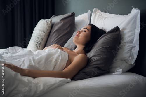Portrait of a young cute brunette girl sleeping on a bed in light livingroom. Her head is on grey pillow and she is covered with white blanket