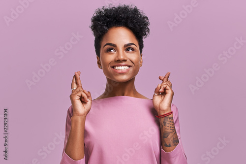 Fototapet Beautiful African American female student crosses fingers with big hope, has positive expression, poses against lavender background