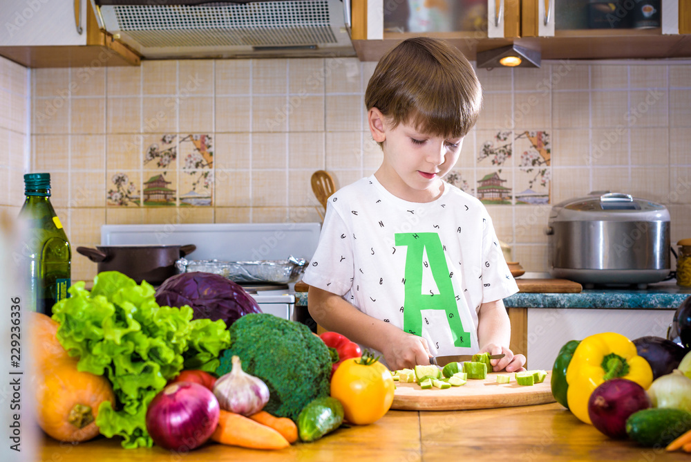 The young boy in cooking standing in the kitchen near table with vegetables The young boy in cooking standing in the kitchen near table with vegetables