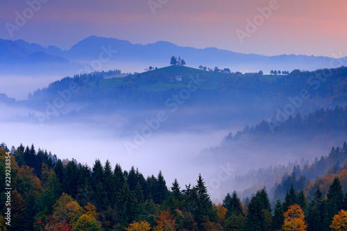 Twilight with fog in Slovenia. Saint Thomas Church, Sveti Tomaz nad Praprotnim, Skofja Loka. Foggy Alps with forest, travel in Slovenia, Europe. Beautiful sunrise with blue sky, autumn nature.