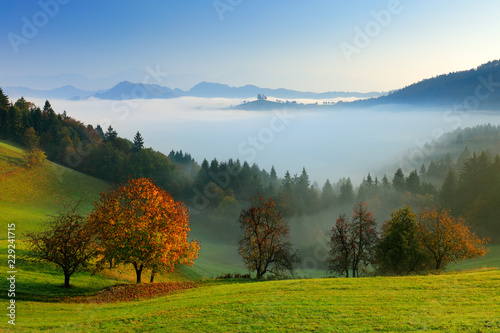 Autumn in Slovenia with fog. Saint Thomas Church, Sveti Tomaz nad Praprotnim, Skofja Loka. Foggy Alps with forest, travel in Slovenia, Europe. Beautiful sunrise with blue sky, green nature. photo