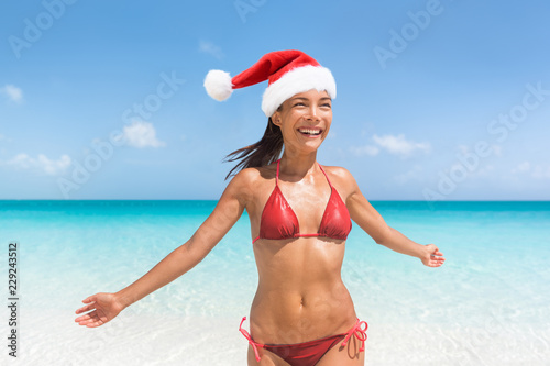 Christmas Caribbean beach tropical getaway Santa Claus woman. Happy Asian bikini girl with slim body for weight loss new year resolution concept. Travel vacation holidays under the tropical sun.
