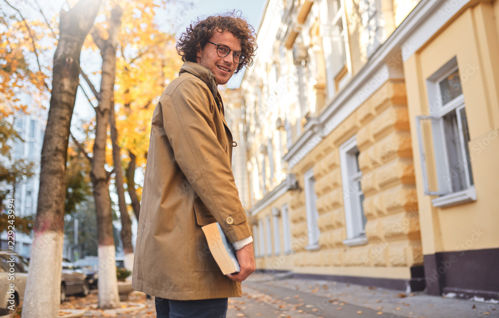 © iuricazac - Rear view of young man with glasses posing with book outdoors. College male student carrying books in campus in autumn street. Smiling guy wears spectacles and curly hair reading books outside