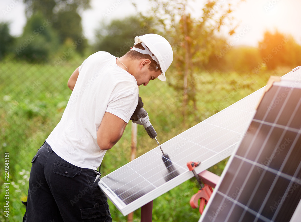 Profile of professional worker in helmet connecting solar photo voltaic panel to metal platform using electrical screwdriver outdoors on bright sunny day on blurred green rural landscape background.