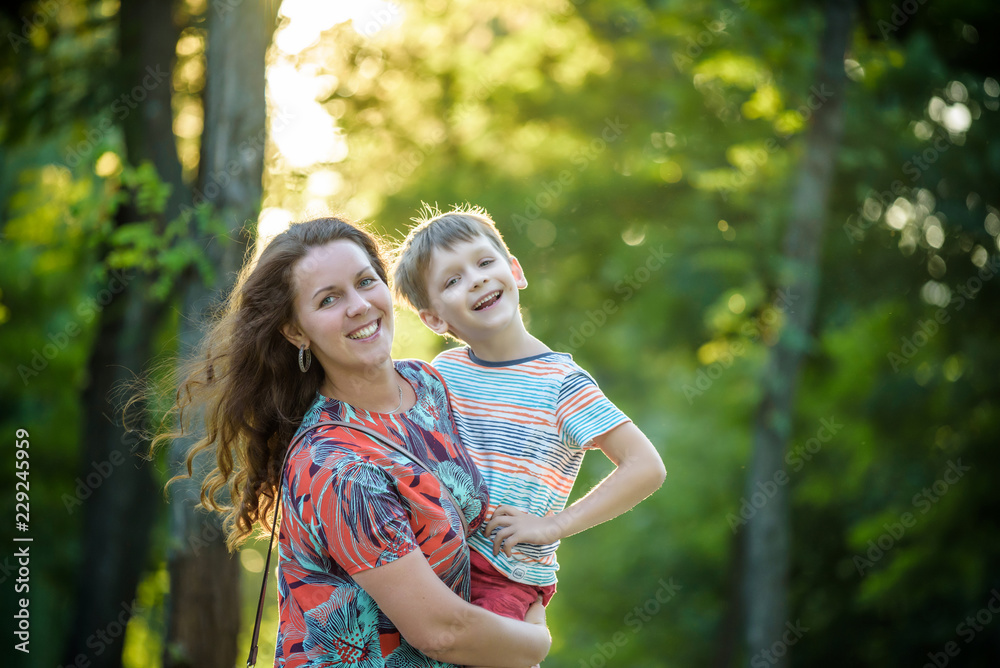 Beautiful woman and her cute little son are hugging and smiling, on nature park background. Boy is looking at camera. Concept of friendship between children and parents, happy family Beautiful woman and her cute little son are hugging and smiling, on nature park background. Boy is looking at camera. Concept of friendship between children and parents, happy family