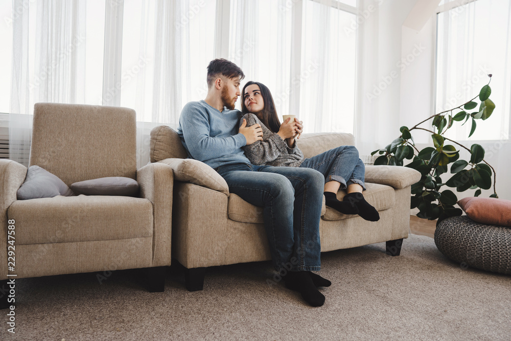 Young couple relaxing at home sitting at sofa with hot drinks. Warming, calmness, home owning, comfort concept