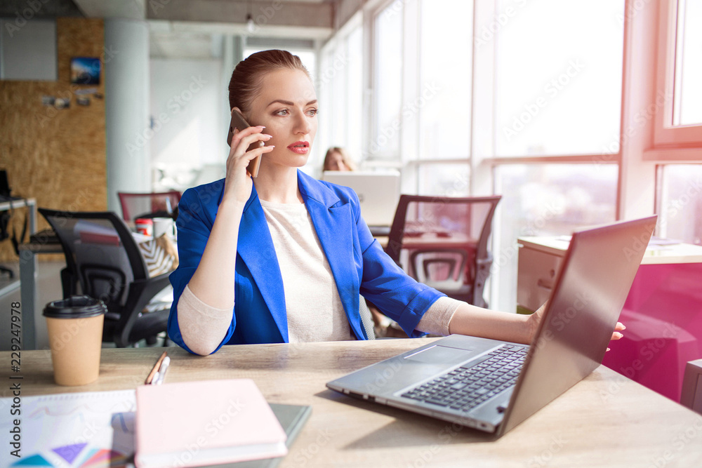 Busy and serious business woman is sitting at the table in office and looking to the laptop. She is working. Also the lady is talking on the phone. Busy and serious business woman is sitting at the table in office and looking to the laptop. She is working. Also the lady is talking on the phone.