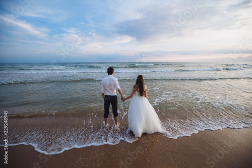A beautiful couple of newlyweds, the bride and groom walking on the beach. Gorgeous sunset and sky. Wedding dresses, a white luxury dress for a girl. Family concept, honeymoon.