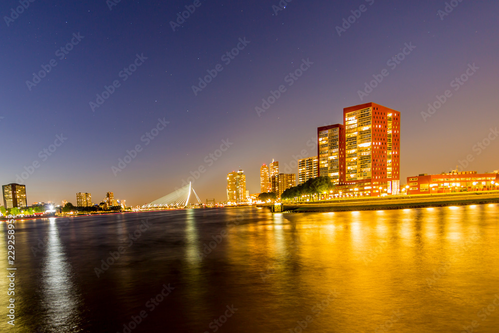 Obraz premium Night cityscape of buildings, yellow lights reflected in the water and the Erasmus cable-stayed bridge over the Maas river in the background in Rotterdam, clear sky in the Netherlands