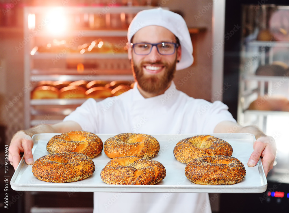 © Evgeniy Kalinovskiy - attractive Baker in white uniform holding a tray with freshly baked bagels with sesame and poppy seeds on the background of a bread factory or bakery