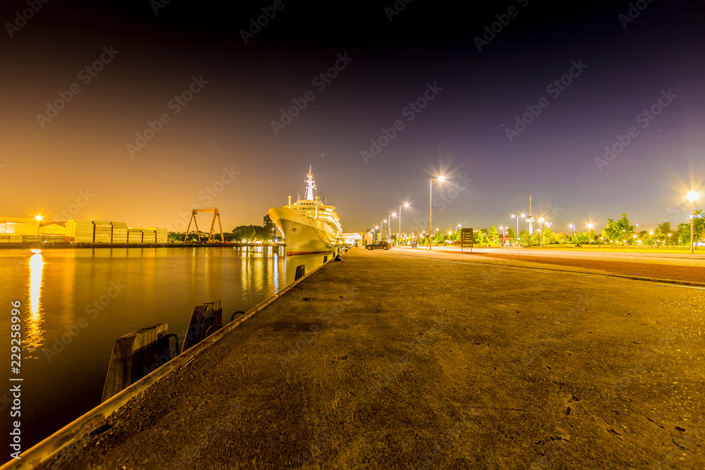 Night view of the port of Rotterdam with a cruise ship anchored on the quay, empty street with illuminated lamp posts and trees in the background, quiet night with a clear sky in the Netherlands