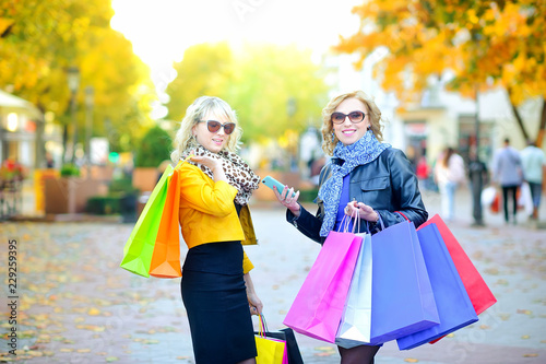 two happy girls with shopping bags and smartphone are smiling while standing on the street