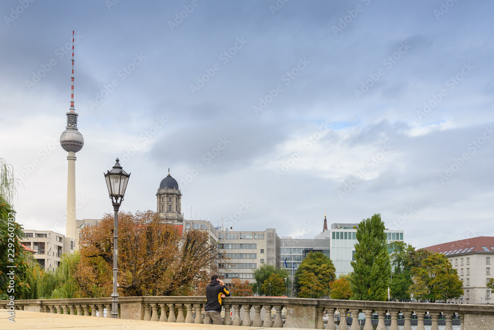 Man standing and talking on the phone at the bridge cross Spree river with background of Berlin cityscape and Television tower at Alexanderplatz during cloudy sky day. 