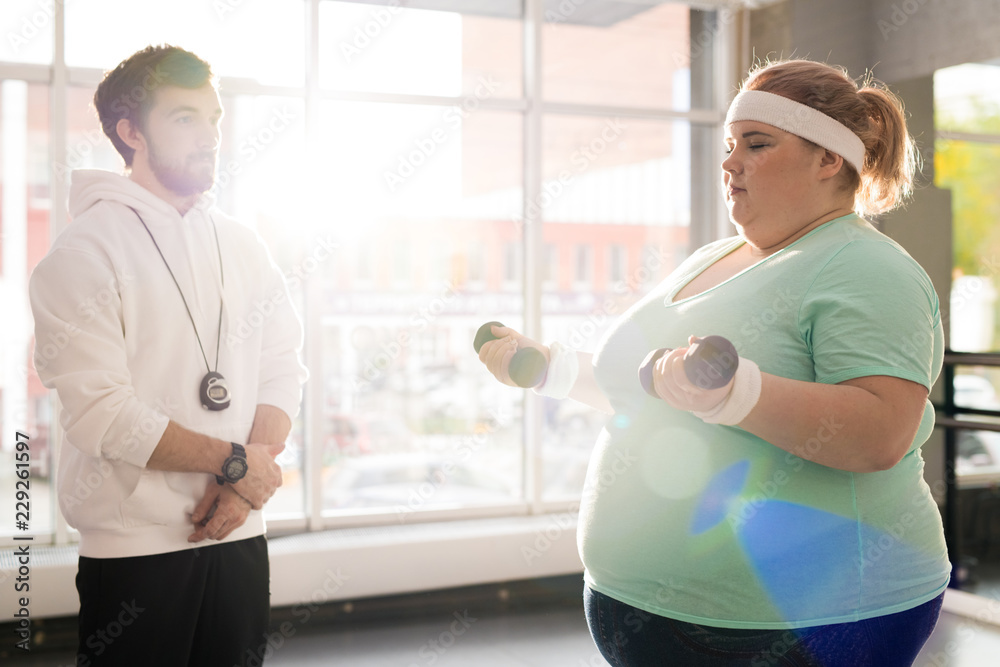 Fototapeta premium Waist up portrait of obese young woman working out with dumbbells during weight loss training with fitness instructor in sunlight