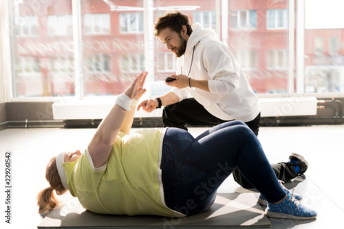 Side view portrait of obese young woman doing crunches during workout with fitness instructor in sunlight, copy space