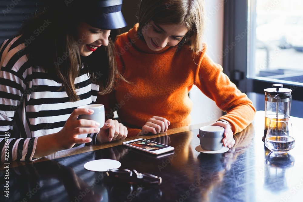 Fototapeta premium Two young women sitting in a cafe, drinking their morning coffee and surfing the net on smart phones.