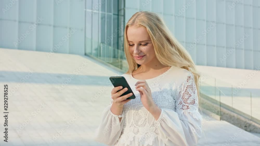 An attractive young blonde lady walking and using a phone in town. Medium shot. Soft focus. Dolly shot An attractive young blonde lady walking and using a phone in town. Medium shot. Soft focus. Dolly shot