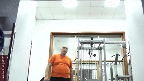 Fat handsome man in orange t-shirt in a gym is going away from a room. Bottom view