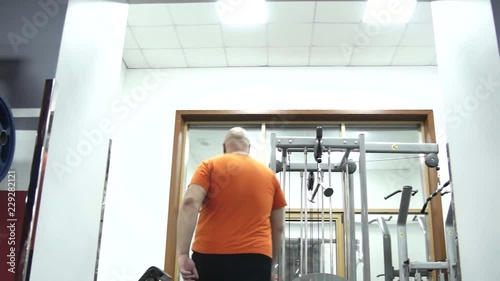 Fat handsome man in a gym in orange t-shirt is coming, the view from the bottom up