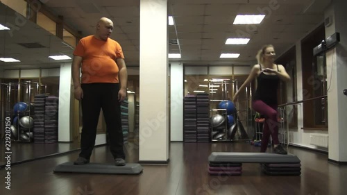Fat funny man is standing on a step and looking with bored at young beautiful woman actively doing step aerobics.
