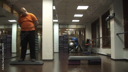 Fat funny man is standing on a step in a fitness room with a confused look, camera moving from right into left