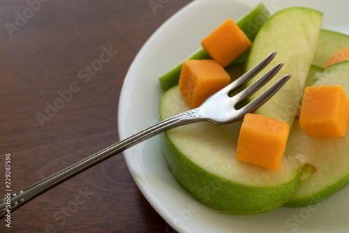 A fork atop cheddar cheese squares with green sliced apples on a white plate atop brown wood table.