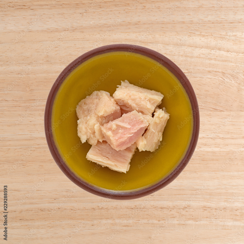 Top view of a small bowl with solid white albacore tuna in olive oil chunks on a wood table. Top view of a small bowl with solid white albacore tuna in olive oil chunks on a wood table.