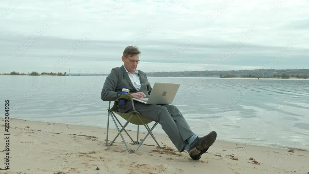Mature businessman sitting and relaxing near a river. Man in suit and using laptop. Mature businessman sitting and relaxing near a river. Man in suit and using laptop.