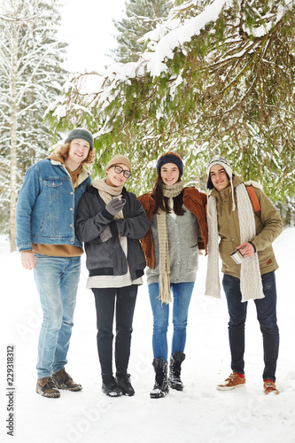 Full length portrait of group of happy young people posing in winter resort standing under fir tree in beautiful snowy forest