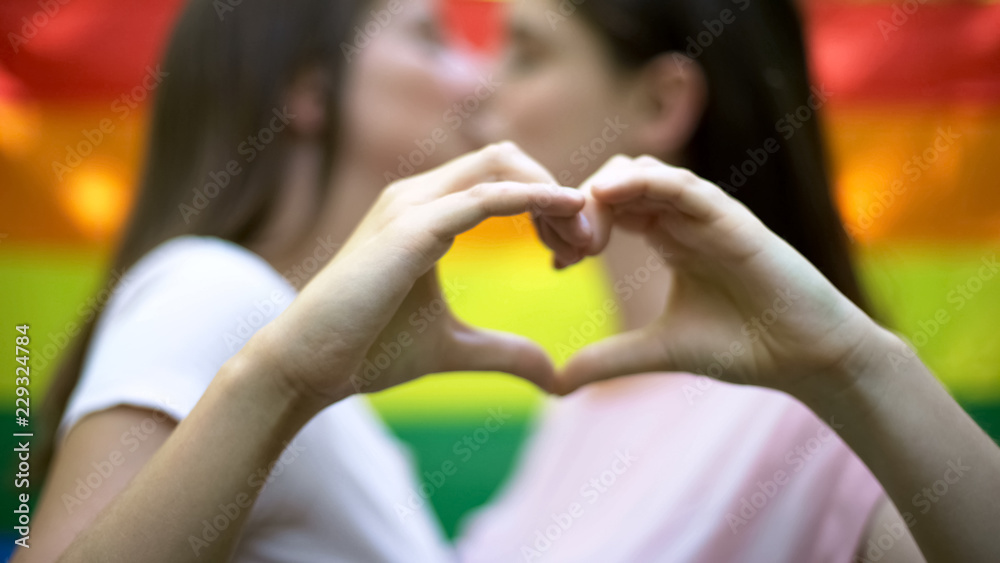 Lesbians showing love sign, kiss against rainbow flag background, minority right