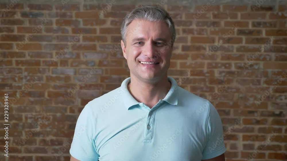 Cute caucasian mature man is showing like sign at camera and smiling while standing isolated in red brick studio