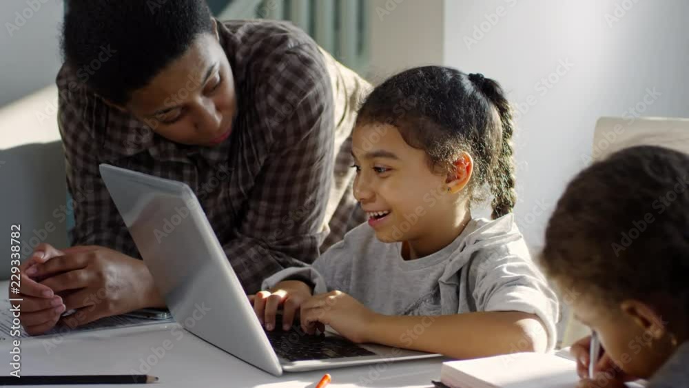 PAN of black woman explaining something to playful primary age daughter typing on laptop and doing homework; cute preschooler sitting beside and writing in notebook